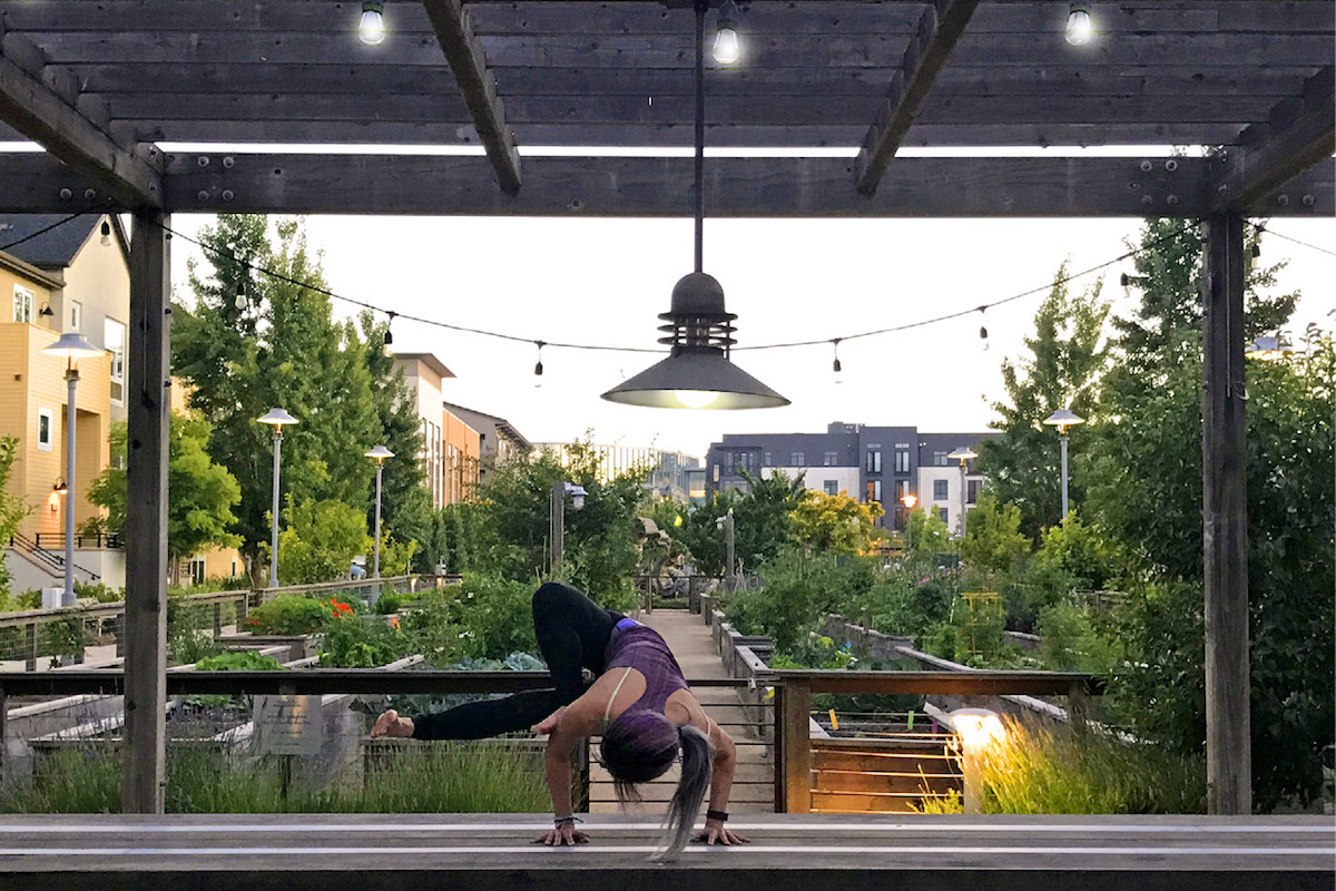 young woman doing yoga in a park with green hedges and trees, townhouses, and wood planked walk ways in the background