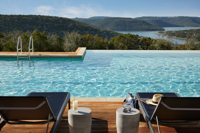 sweeping view from pool terrace of lush green mountains and a blue river. Two black patio recliners sit on maple colored wood decking and are before turquoise pool water