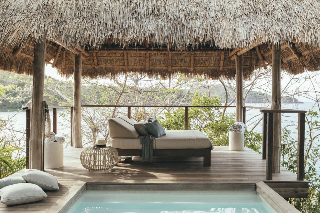 island hut with luxury chaise lounge below it. white upholstered cushions and grey throw pillows. small jacuzzi in the foreground with a thatched roof and the ocean in the background