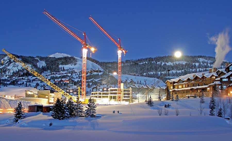 wintertime construction site with large cranes and log condo building structures under construction with tall mountain range directly behind it - nigh time image with industrial lights illuminating the site, and a bright, full moon above