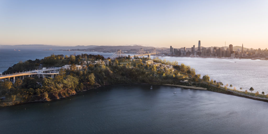 aerial views of yerba buena island located in san francisco bay. the image features an island connected to by a tall bridge in the middle of the ocean with a city view behind it and mix-use residential building stepped into the island's mountainous terrain
