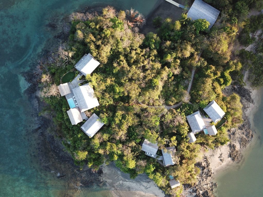 aerial image of a private island with small, hatched roofs nestled amongst lush jungle greenery and surrounded by blue ocean water