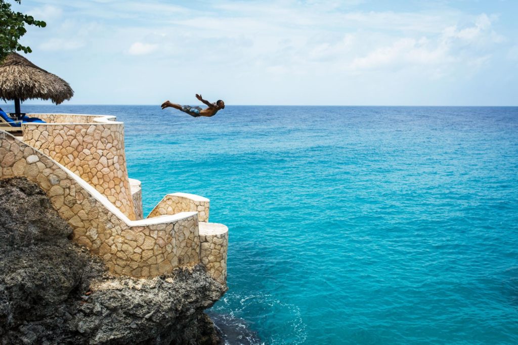 a man cliff diving from a stone wall into clear, crystal blue ocean water in jamaica