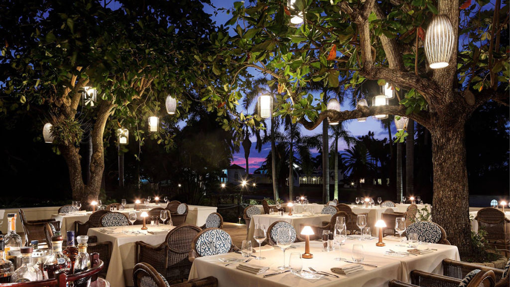 outdoor dining in a beach setting with large trees and lanterns hanging from them to overhead light dining tables with rattan chairs and blue and white upholstered back rests