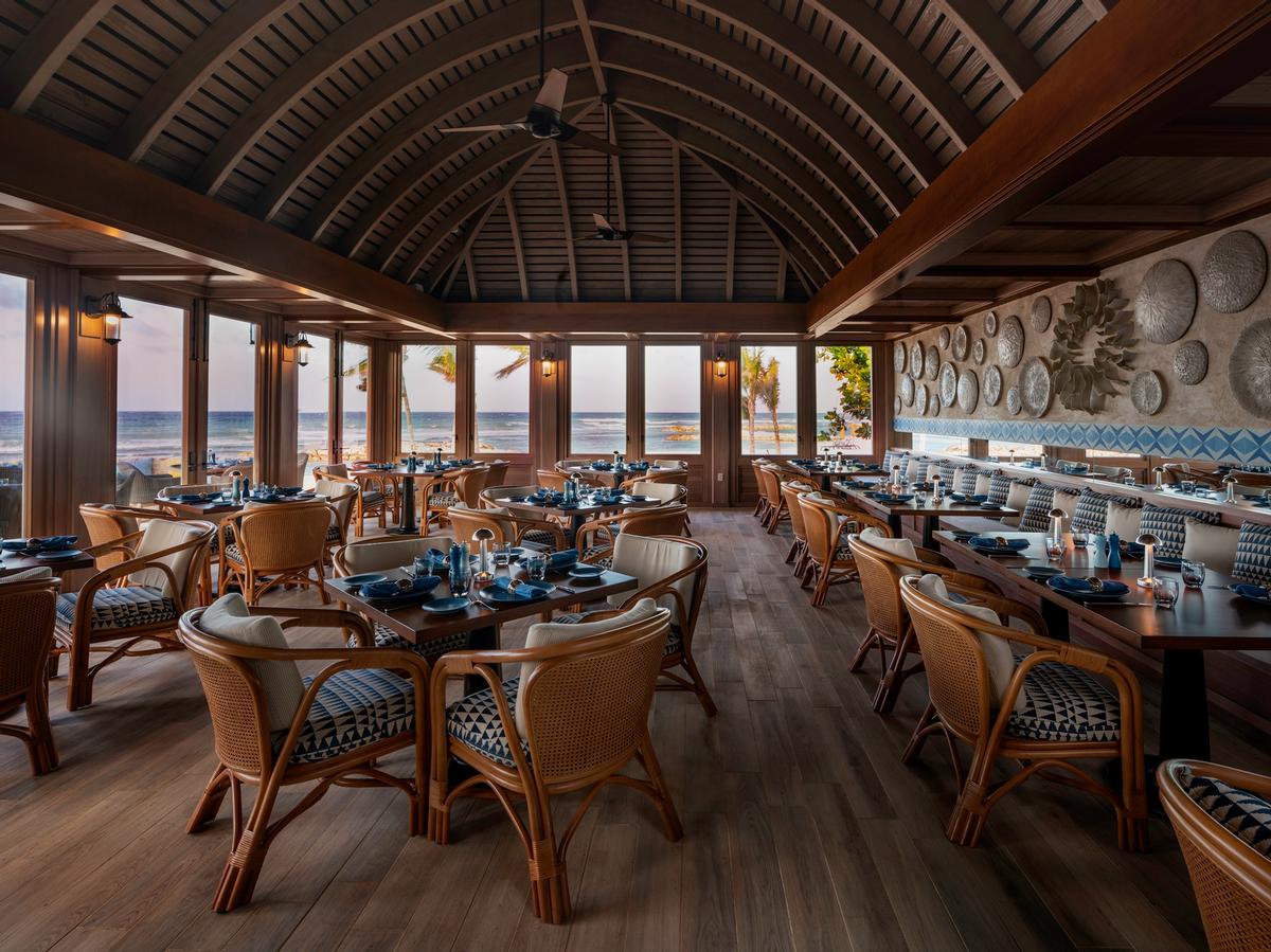 waterfront restaurant with dark wood paneling on the ceiling that mimics that of an upside down or interior of a canoe. A custom 3 dimensional mural hands on the lateral wall with sea shell like designs. Blue accents on rataan upholstered dining chairs and blue tiles contrast the brown and wood palatte,