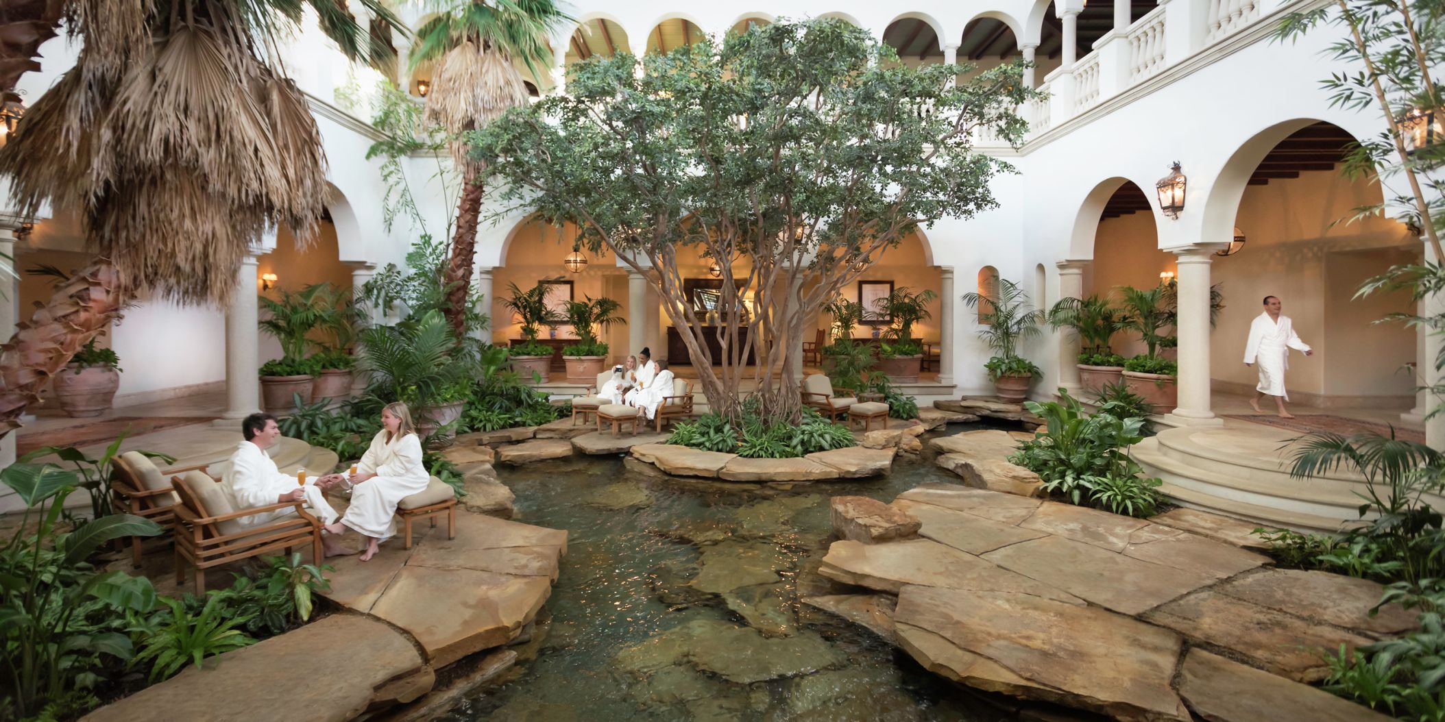 Spa atrium with classic, addison mizner style inspired architecture with lush, think greenery and people with spa robes waiting for their treatments.