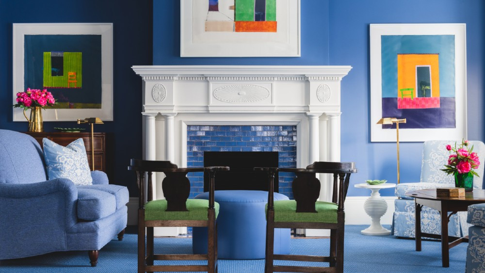 blue guest welcome area at a small boutique like hotel with a blue tile fireplace and a white mantle. blue overstuffed furniture adorns the room with modern art hanging on blue walls.