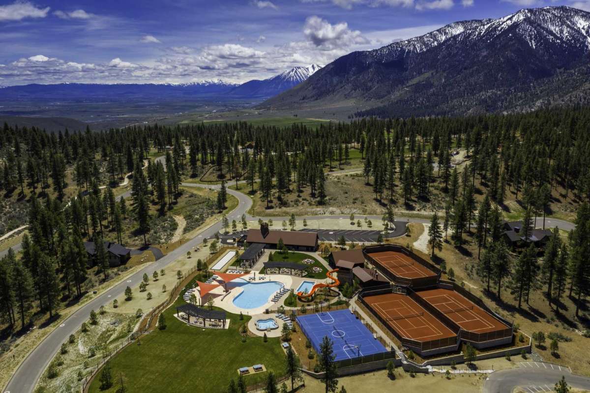 aerial photo of lake tahoe mountains with a camp in the middle - a pool and tennis courts surround the main cabin structure. lush evergreen trees and snow capped mountains under a blue sky surround the site.