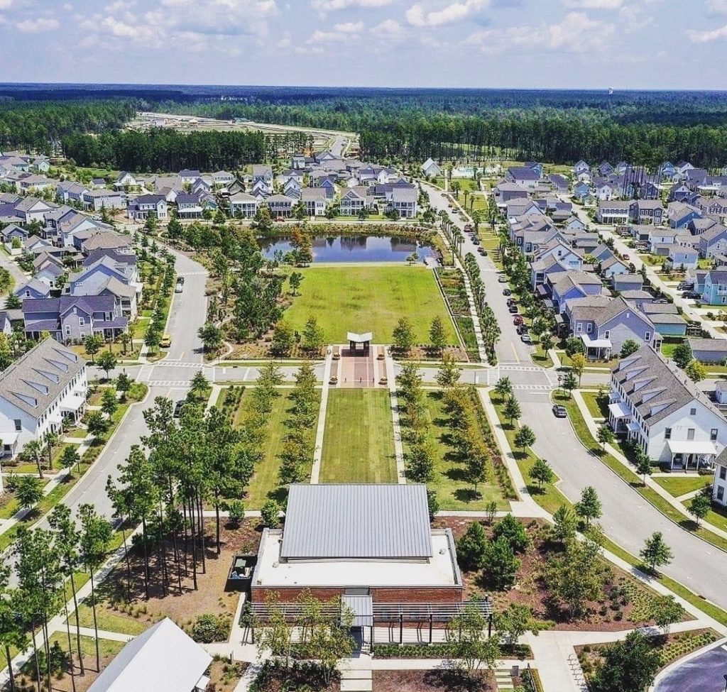 aerial view of a planned community centered around a green lawn and small lake with a clubhouse at the top and houses surrounding with lush greenery in the background