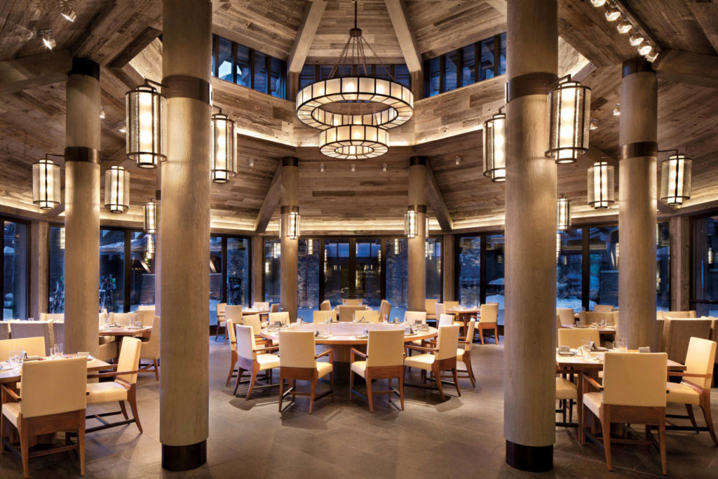 interior view of a large log cabin inspired hotel restaurant, large, floor to ceiling glass windows look out onto the rocky mountains where snow caps the landscape. Comfy dining chairs and large, modern chandeliers adorn the dining room space