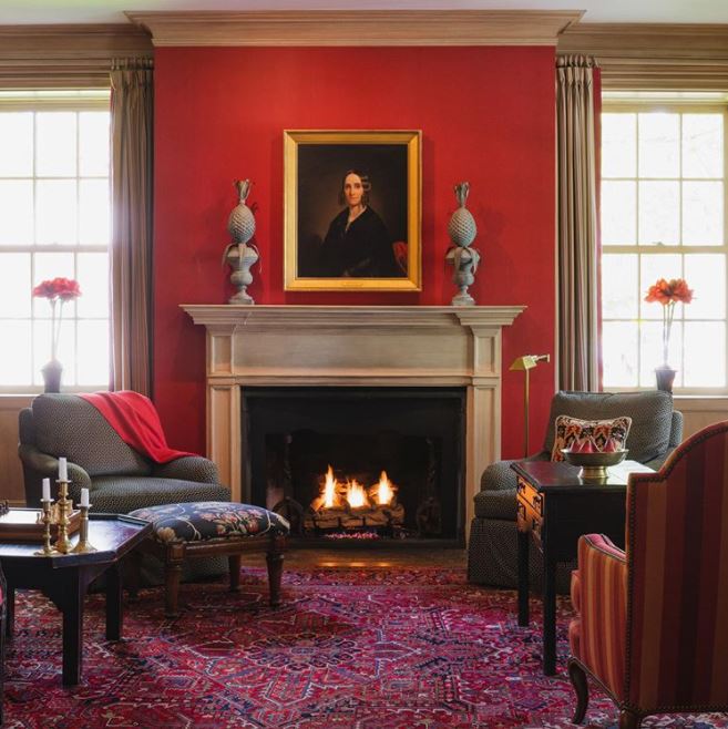 interior shot of a living room like space with a red accent wall and fine art portrait of a woman. purple and red patterned carpeting is on the floor and atop of it is several pieces of transitional style club chairs in charcoal and reds. A large fireplace is at the center focal point of the photo