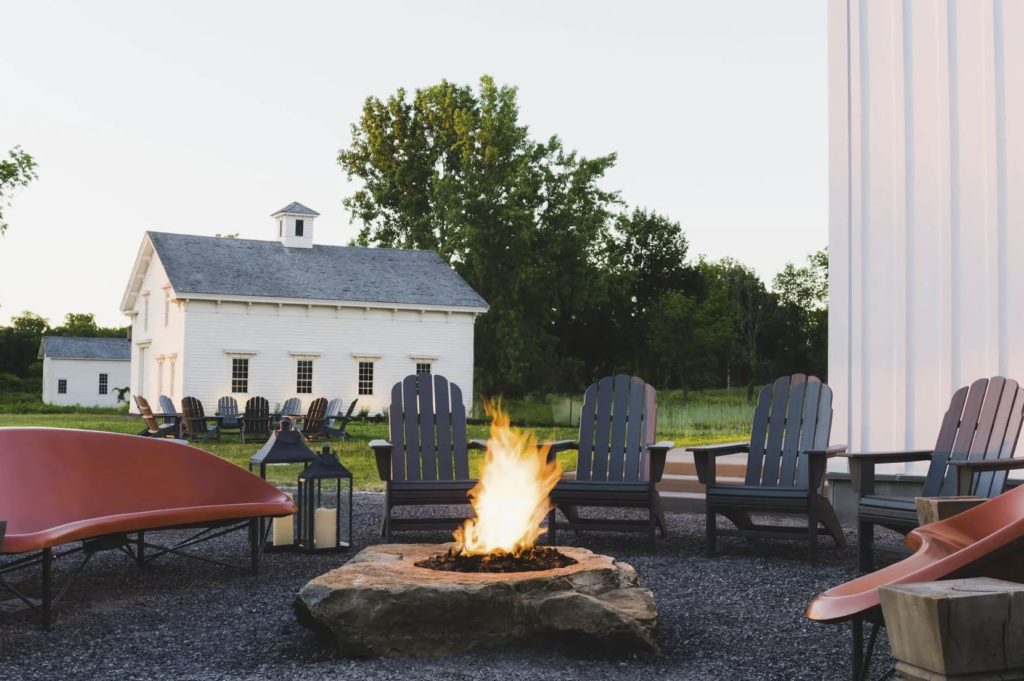 outdoor fire pit on black rocks with windsor chairs and red modern seating surrounded by white barn structures