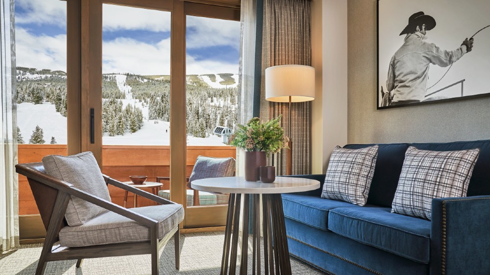 interior rendering of a guest room looking out over a large snowy mountain top with a chair lift in the far distance. The guest room has a blue, modern love seat with two throw pillows, a modern, wood accented arm chair and a contemporary bronze and marble side table. A black and white photo of a man with herding cattle and wearing a cowboy hat with his back to us is on the center of the back wall above the love seat
