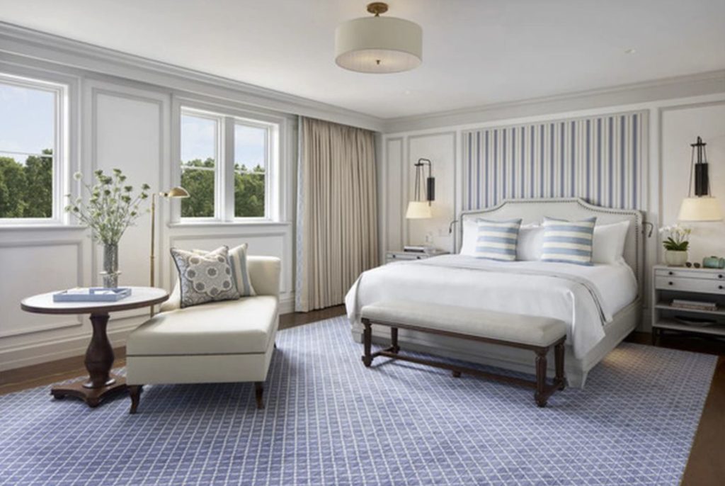 photo of a guest room with blue accents a large white linen covered bed and a chaise. LArge windows with a view of greenery are on the left hand wall