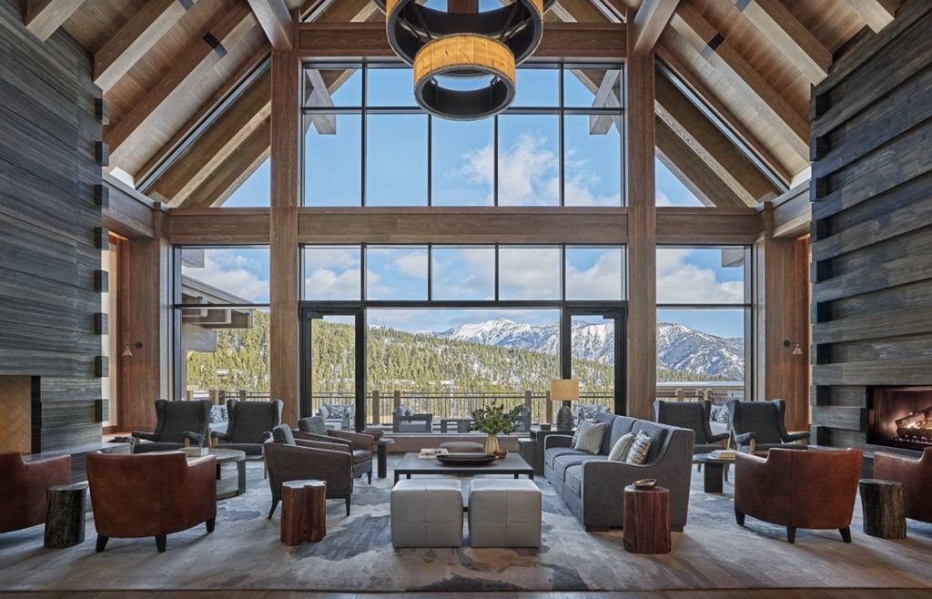 interior view of a large log cabin inspired hotel, large, floor to ceiling glass windows look out onto the rocky mountains where snow caps the landscape. Comfy club chairs and large, modern chandeliers adorn the living room space