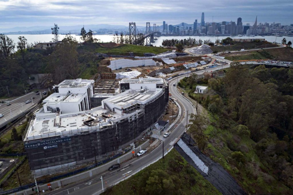 aerial of a construction site with a view of san francisco behind it. A large condo building is nestled into the hillside and is currently under construction.
