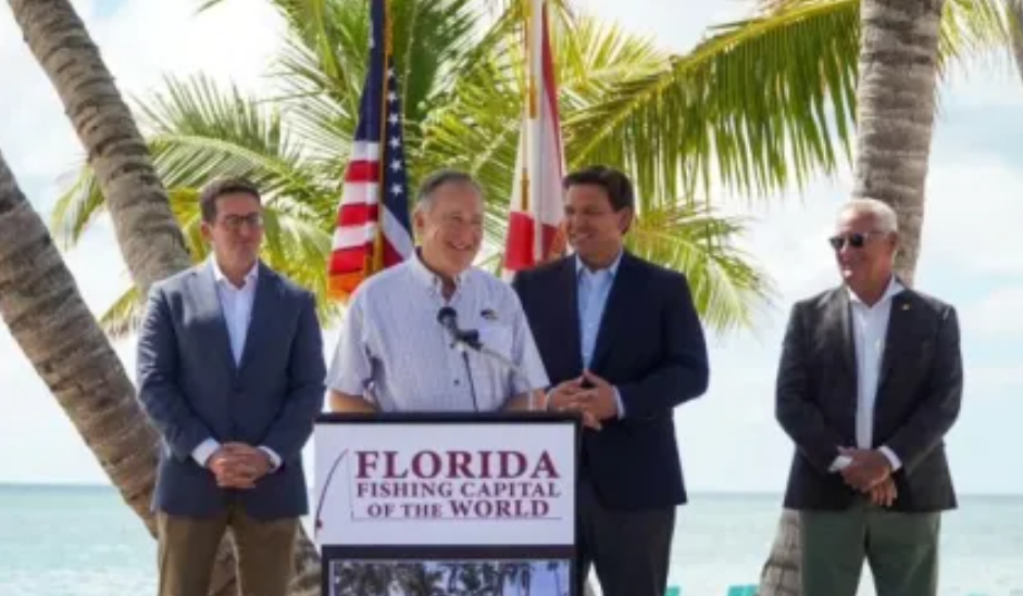four men on stage during a press announcement that overlooks the ocean with palm trees in the background