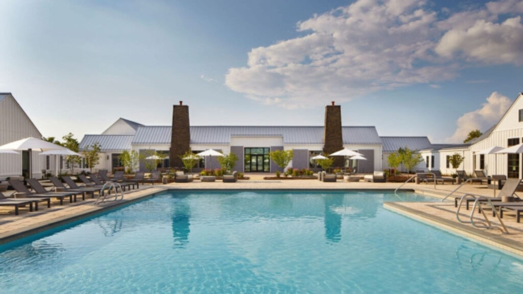 photo of a pool deck with luxe recliners, umbrellas and landscaping dotted around the pool. A barn like structure lies at the far end of the pool.